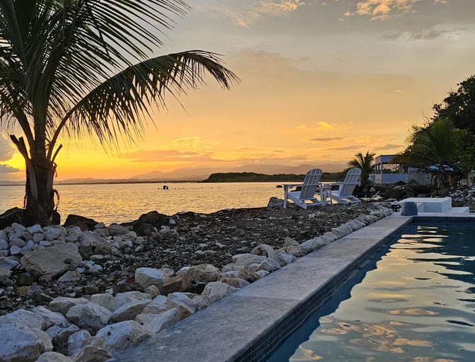 Beachfront pool and lounge chairs at sunset with palm tree, rocky shore, and calm ocean beyond